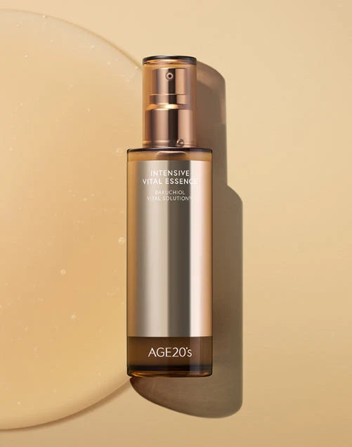 Two bottles of AGE 20 INTENSIVE VITAL ESSENCE, one standing upright and the other lying on its side, are displayed on a beige surface with two small liquid drips. Available to buy at MyAllures.