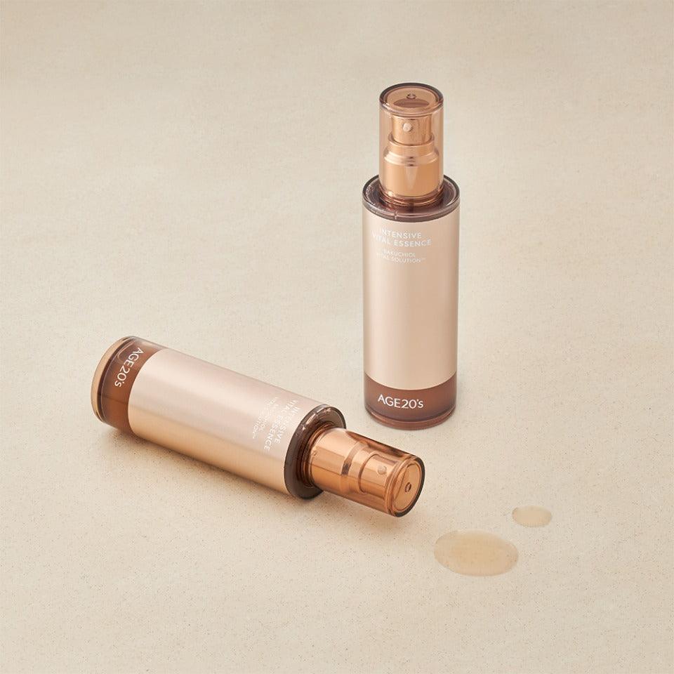 Two bottles of AGE 20 INTENSIVE VITAL ESSENCE, one standing upright and the other lying on its side, are displayed on a beige surface with two small liquid drips. Available to buy at MyAllures.