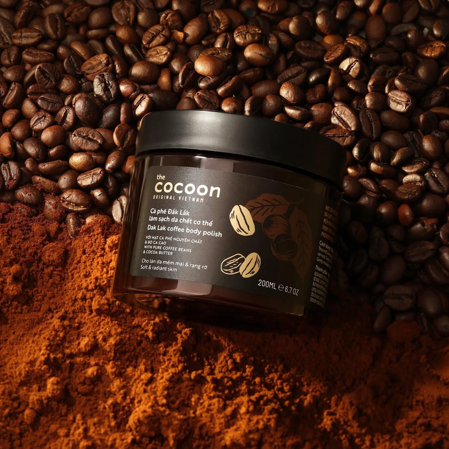 Hands holding an open jar of COCOON DALAK COFFEE BODY POLISH and a handful of coffee beans against a beige background, available to buy at MyAllures.