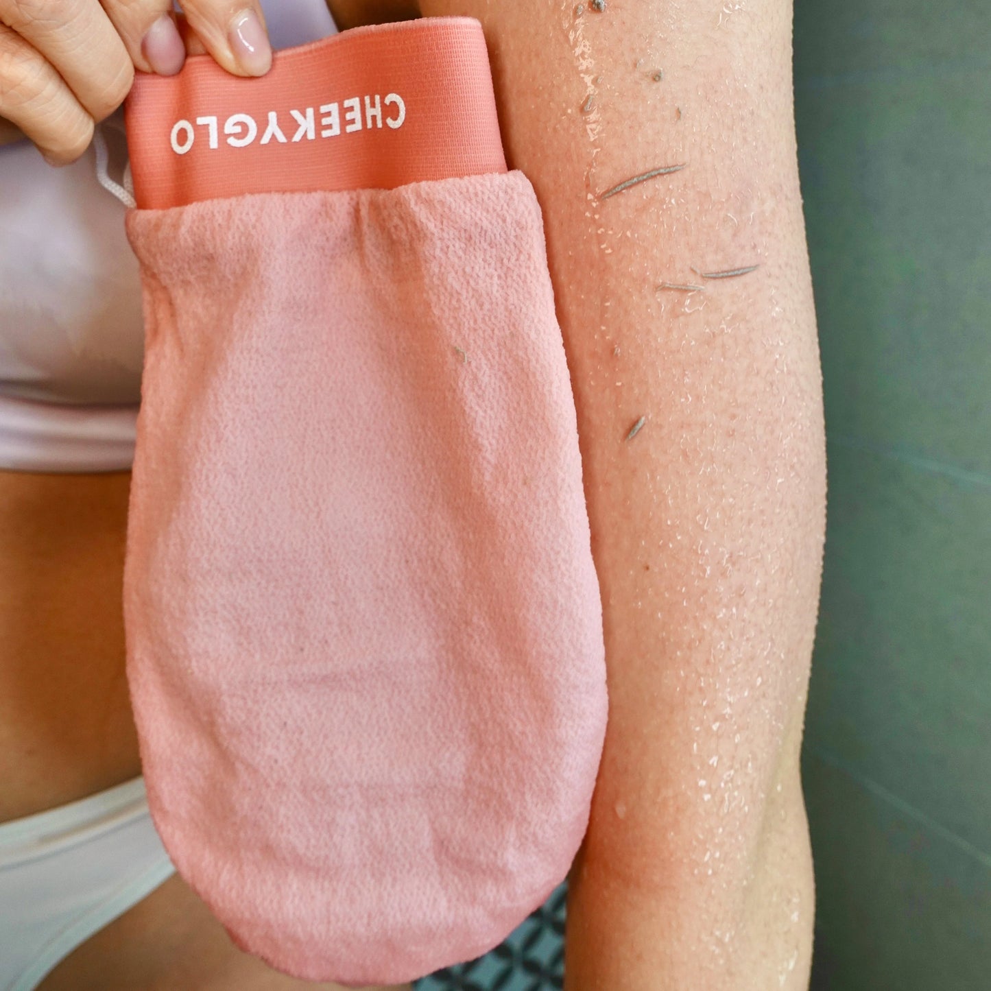 A person wearing white bikini bottoms and the CHEEKYGLO EXFOLIATING LOOFAH GLOVE with peach-colored ruffles against a pink background—find this look at MyAllures, brought to you by CHEEKYGLO.