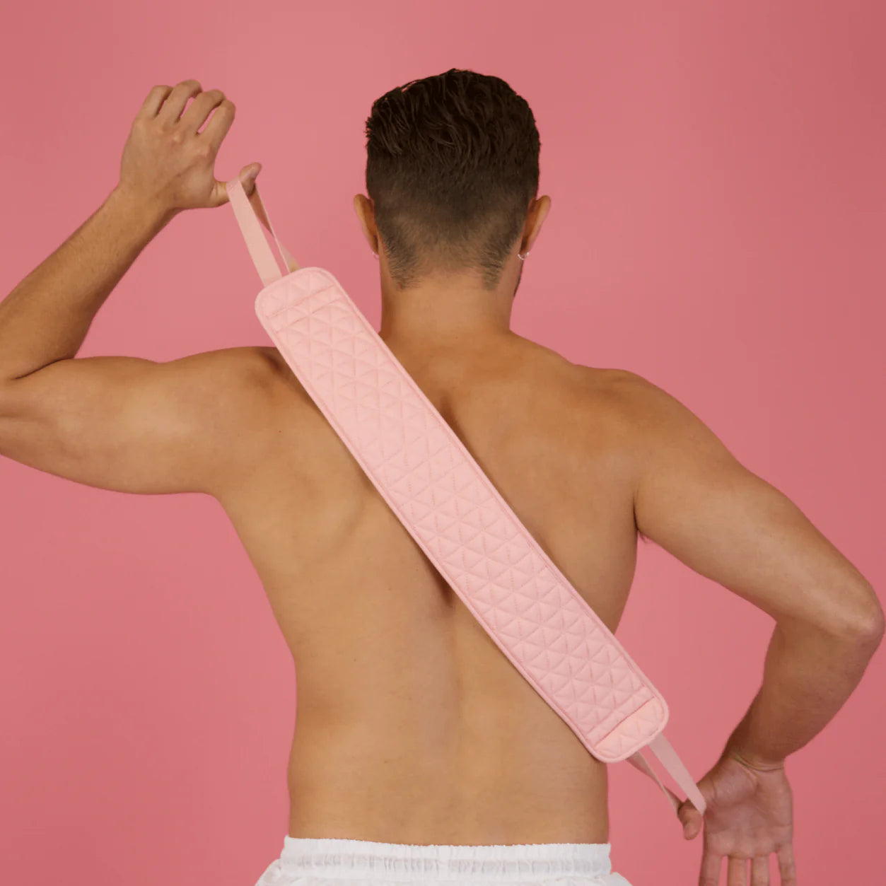 A woman in white underwear uses a CHEEKYGLO EXFOLIATING BACK SCRUBBER against a pink background. Buy at MyAllures.