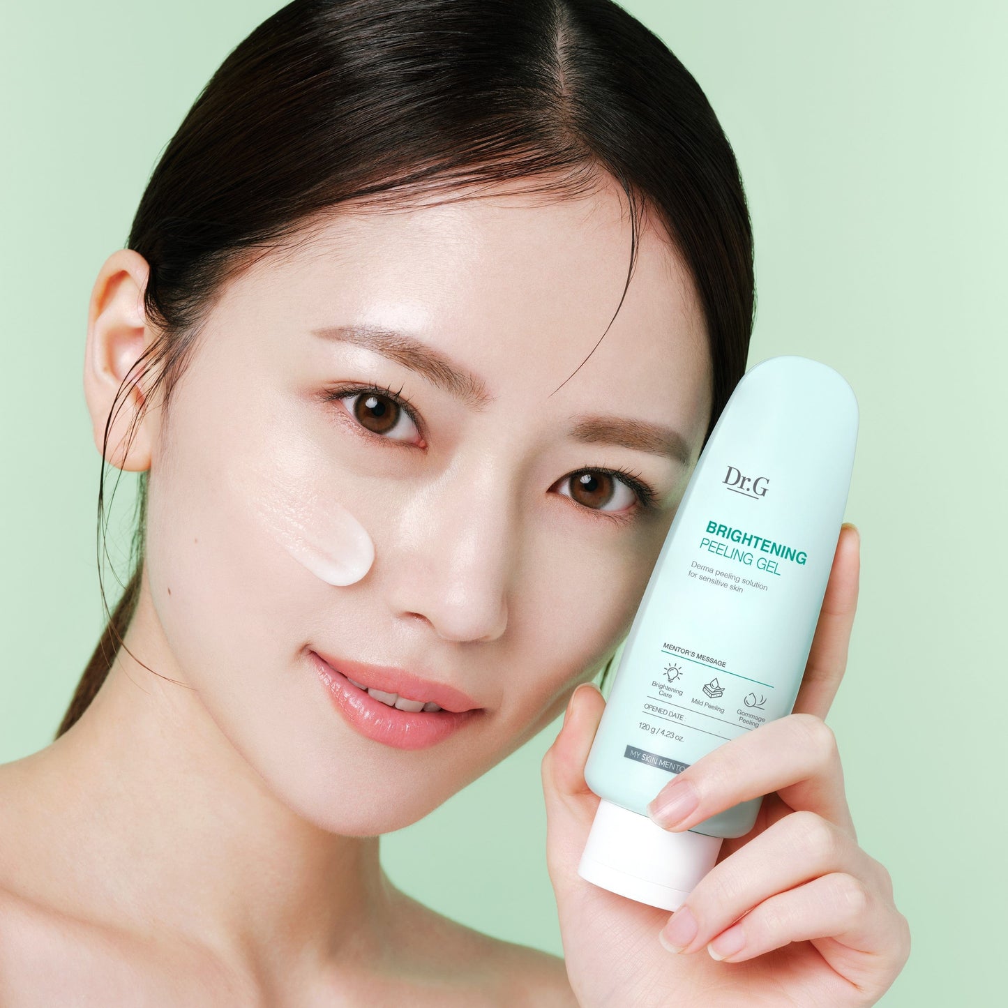 A woman holds DR.G BRIGHTENING PEELING GEL next to her face, showing a small amount of the product applied on her cheek. Purchase now at MyAllures.