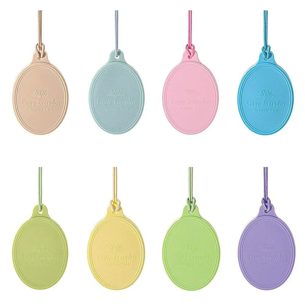 Five oval-shaped leather key holders in pink, mint, red, green, and peach colors are arranged in open gift boxes on a white fabric background. A card with the text "love is home" from the ENJOY EVERYDAY AIR FRESHENER SCENTED CARD collection by ENJOY EVERYDAY is partially visible. Available to buy at MyAllures.