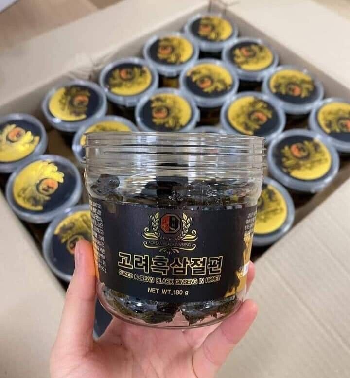 A hand holds a piece of KOREAN SLICED BLACK GINSENG HONEY, characterized by its dark, wrinkled appearance. In the background, several small containers with yellow and black lids are visible, available to buy at MyAllures.