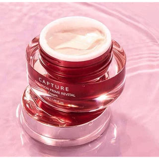 A red and pink jar labeled "AHC Cature Solution Prime Revital Cream," filled with luxurious cream, is placed on a reflective pink surface, with the lid removed and resting beside it. Discover this premium cream by AHC and purchase it at MyAllures.