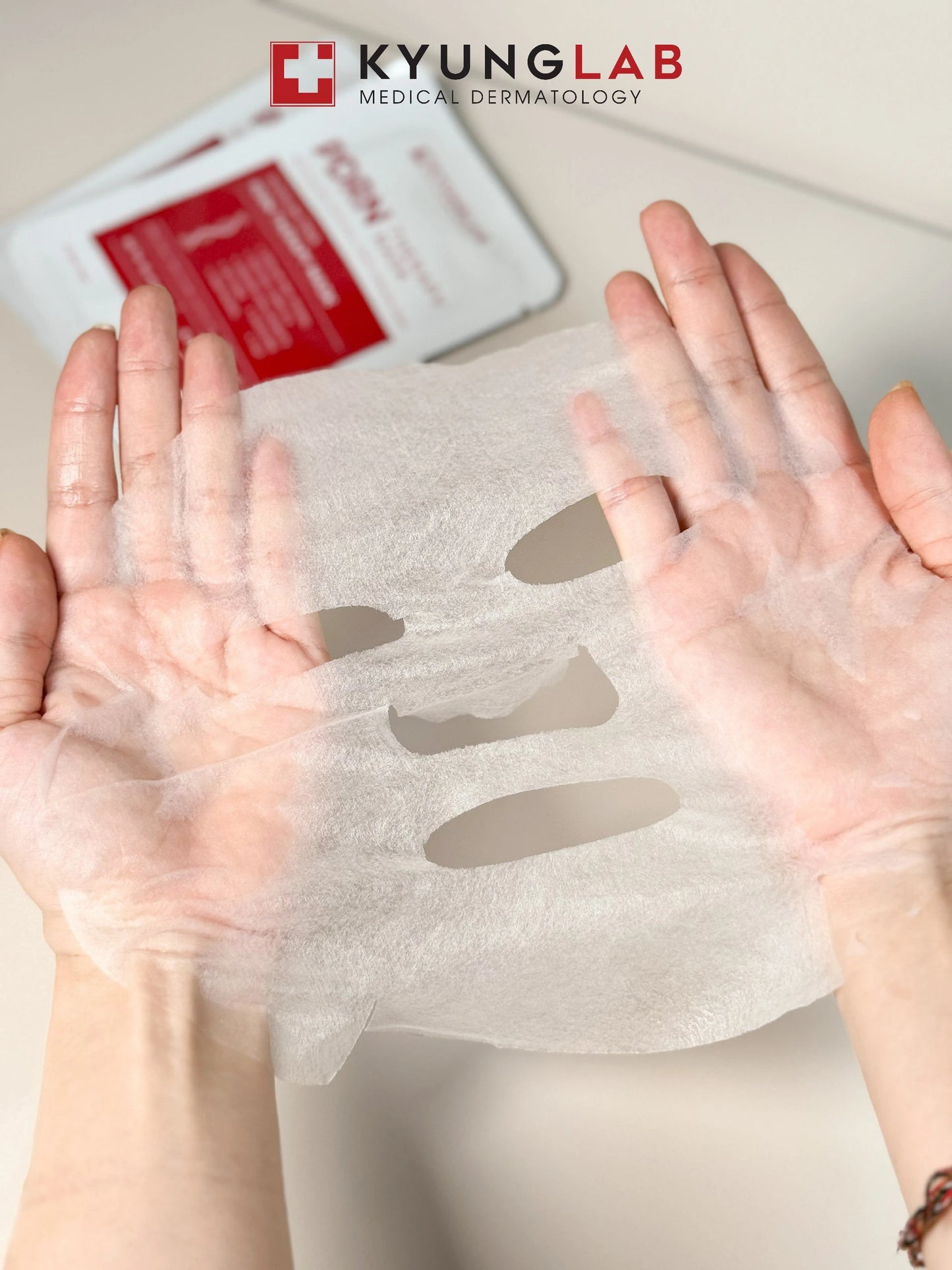 Hands holding a translucent KYUNGLAB PDRN THERAPY MASK with eye, nose, and mouth cutouts. A brochure and the name "KYUNGLAB Medical Dermatology" are visible in the background. You can buy it at MyAllures for premium skincare solutions.